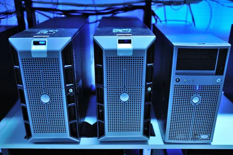 Three silver computer servers with perforated front panels on a white shelf under blue lighting.