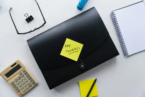 The modern tools of accounting (irony)- Black envelope with a sticky note saying Pay Taxes surrounded by calculator, glasses, notebook, and pen on desk -