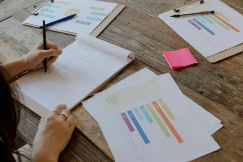 Person writing notes on a notepad with project charts and colorful bar graphs on wooden table