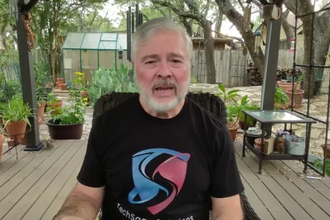 Mature man with gray hair and beard sitting on a wooden patio surrounded by potted plants wearing a TechSoup Solutions t-shirt.