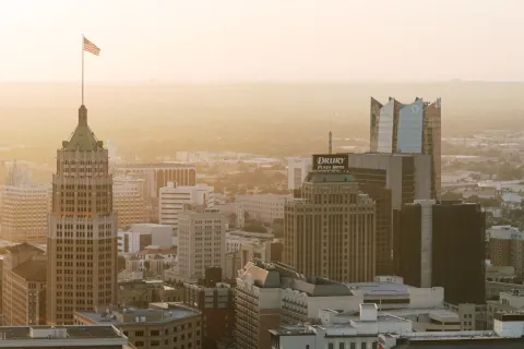 Downtown cityscape at sunrise with tall buildings, including a tower with a waving American flag.
