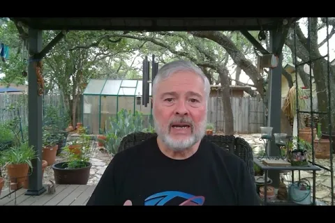 Older man with gray beard speaking outdoors on a covered patio surrounded by plants and garden items.