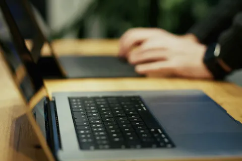 Close-up of a laptop keyboard on a wooden desk with blurred hands typing on another laptop.
