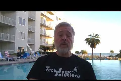 Man with gray hair and beard wearing a Tech Sage Solutions T-shirt by a poolside at a resort near palm trees and ocean.