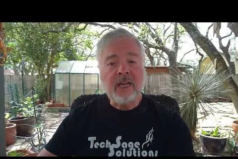 Older man with gray beard wearing Tech Sage Solutions shirt sitting outdoors in a garden with plants and greenhouse.
