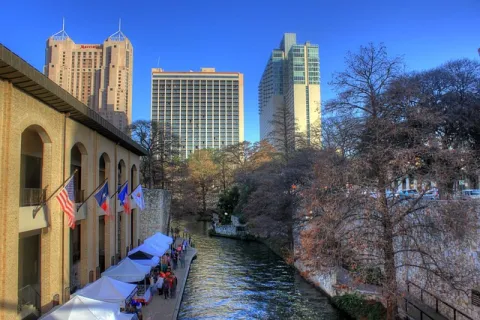 San Antonio Riverwalk with flags, tents, and city skyscrapers under a clear blue sky