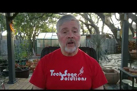 Man wearing a red Tech Sage Solutions t-shirt speaking outdoors in a garden setting with plants and trees.