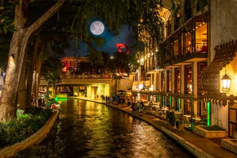 Night view of a lively riverwalk with illuminated restaurants, a full moon, and reflections in the water