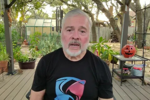 Man with gray beard wearing black t-shirt sitting on a wooden deck with plants and Halloween pumpkin in background
