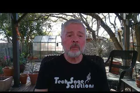 Man with gray beard wearing Tech Sage Solutions shirt sitting outdoors with greenhouse and trees in background