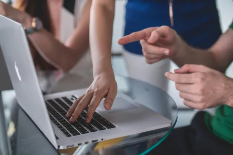 Hands collaborating on MacBook laptop keyboard over glass table in a casual teamwork setting.