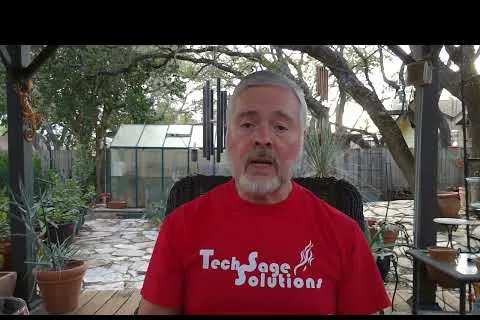 Man wearing red Tech Sage Solutions shirt sitting outdoors with plants and a greenhouse in the background
