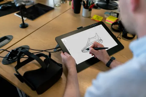 Designer sketching a shoe on a digital tablet using a stylus in a modern workspace with VR headset nearby
