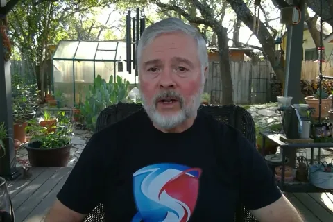 Older man with white beard speaking outdoors on a deck surrounded by plants and trees during daytime.