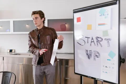 Young man in brown leather jacket presenting startup ideas on whiteboard with notes and charts in office.