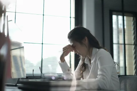 Stressed woman in white shirt sitting at a desk with hand on forehead in a bright office with large windows.
