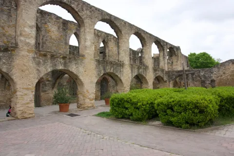Historic stone arches and green bushes in a courtyard under a cloudy sky at an old mission site.