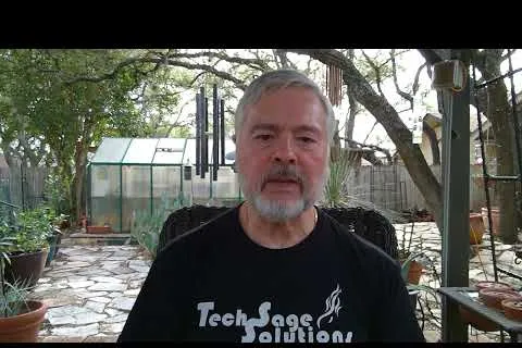 Man with gray beard wearing black TechSage Solutions shirt sitting in a shaded garden patio with greenhouse in background