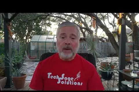 Man with gray hair and beard wearing red TechSage Solutions t-shirt sitting outdoors on a wooden patio with plants.
