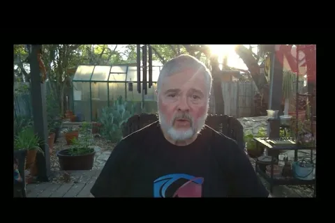 Older man with gray beard in black shirt sitting outside on patio with greenhouse and plants in background during sunset.