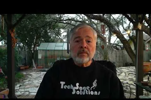 Older man with beard wearing a Tech Sage Solutions shirt sitting outdoors in a patio with trees and a greenhouse.