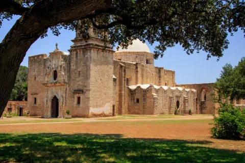 Historic stone mission building with dome and arched entrance under a large tree on a sunny day.