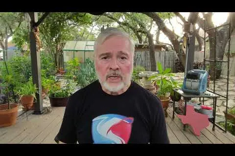 Older man with gray hair and beard wearing a black shirt sitting outdoors on a wooden deck with plants and trees.
