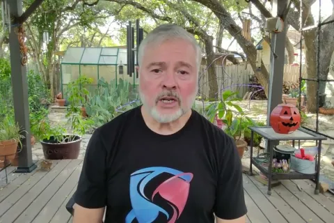 Older man with gray beard wearing a black t-shirt sitting on a wooden deck surrounded by potted plants and Halloween pumpkin decor.