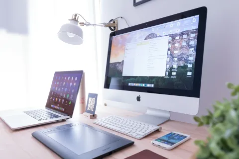 Modern workspace with Apple iMac, MacBook, graphics tablet, smartphone, white keyboard, and desk lamp in bright room.
