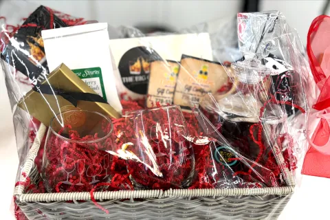 Gift basket wrapped in clear plastic with red shredded paper, glasses, snacks, and a bottle with red ribbon