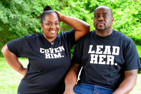 Smiling woman and serious man wearing matching black shirts with Cover Him and Lead Her text outdoors.