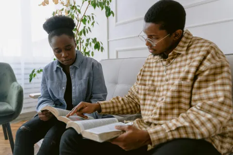 Two individuals reading a book together in a cozy, well-lit living room setting.