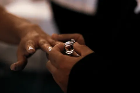Close-up of a person holding a diamond engagement ring near another person's outstretched hand in dim light.