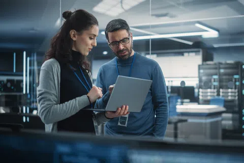 Two IT professionals discuss data on a laptop inside a modern server room with racks and blue lighting.