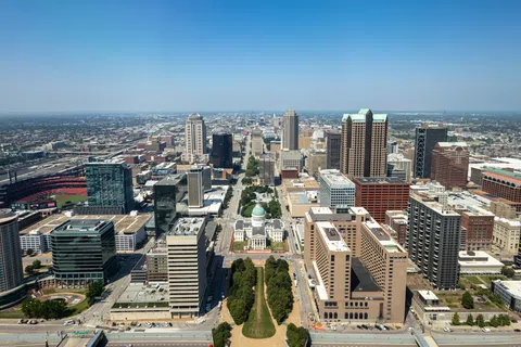 Aerial view of downtown St. Louis with the Old Courthouse, skyscrapers, and sports stadium under a clear blue sky.