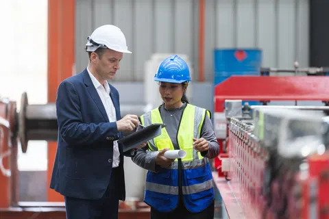 Male engineer in suit and female worker in safety gear discussing plans inside an industrial factory setting