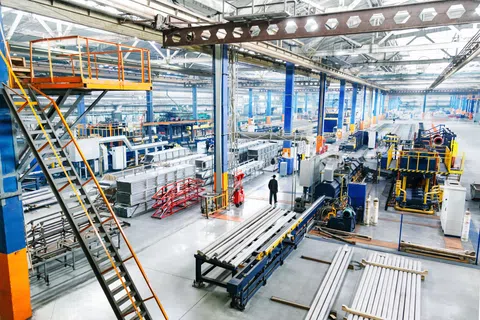 Large industrial factory with metal pipes, machinery, and workers under bright overhead lighting and steel beams.