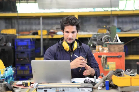 Young engineer working on laptop in an industrial workshop with safety headphones around his neck