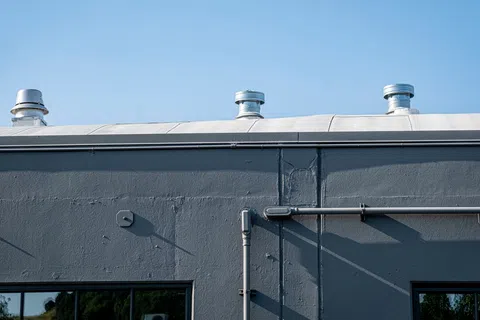 Gray concrete building wall with three ventilation ducts on roof under clear blue sky.