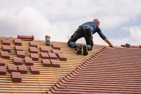 Worker installing brown roof tiles on a wooden roof frame under a cloudy sky.