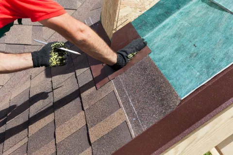 Close-up of a worker installing brown roof shingles with gloves and a pencil marking alignment on a sunny day.