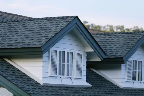 Close-up of house roof with dark gray shingles and two white dormer windows with shutters