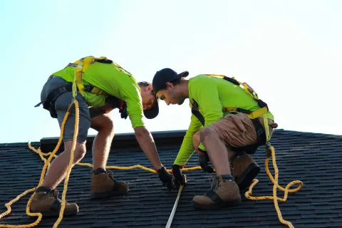 Two roofers in safety harnesses measuring a roof on a sunny day wearing neon green shirts and work boots.