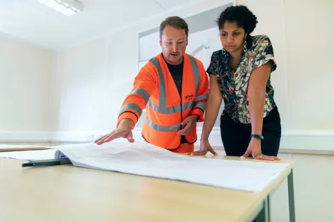 Engineer in orange safety vest explains blueprints to woman in office setting focused on project details.
