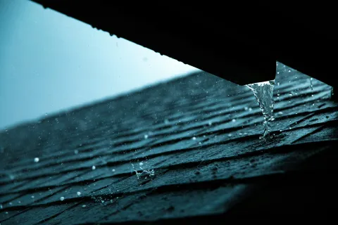 Water dripping from a roof edge onto shingles during rain in moody blue lighting.