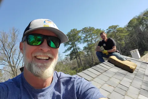 Two men smiling on a rooftop surrounded by trees and clear blue sky on a sunny day