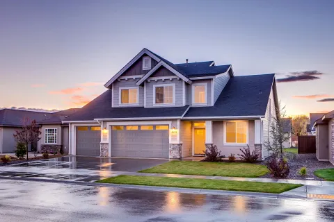Modern two-story suburban house with triple garage and front yard at sunset with wet driveway reflecting light.