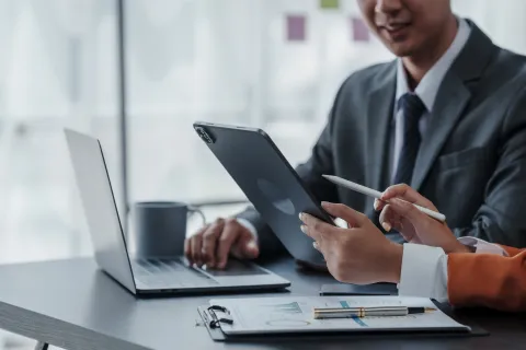 Two professionals collaborating with a tablet and laptop over business charts on a desk in a modern office.