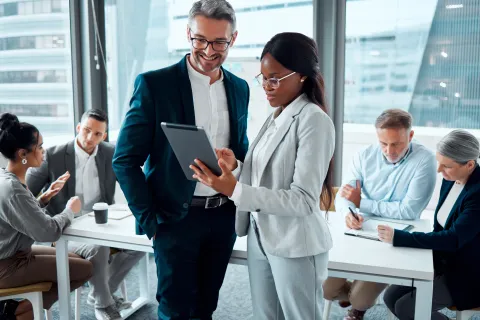 Diverse business team collaborating in modern office, two professionals reviewing tablet while others discuss and take notes.