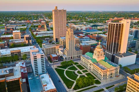Aerial view of a city downtown with historic courthouse, green park, and tall modern buildings at sunset.